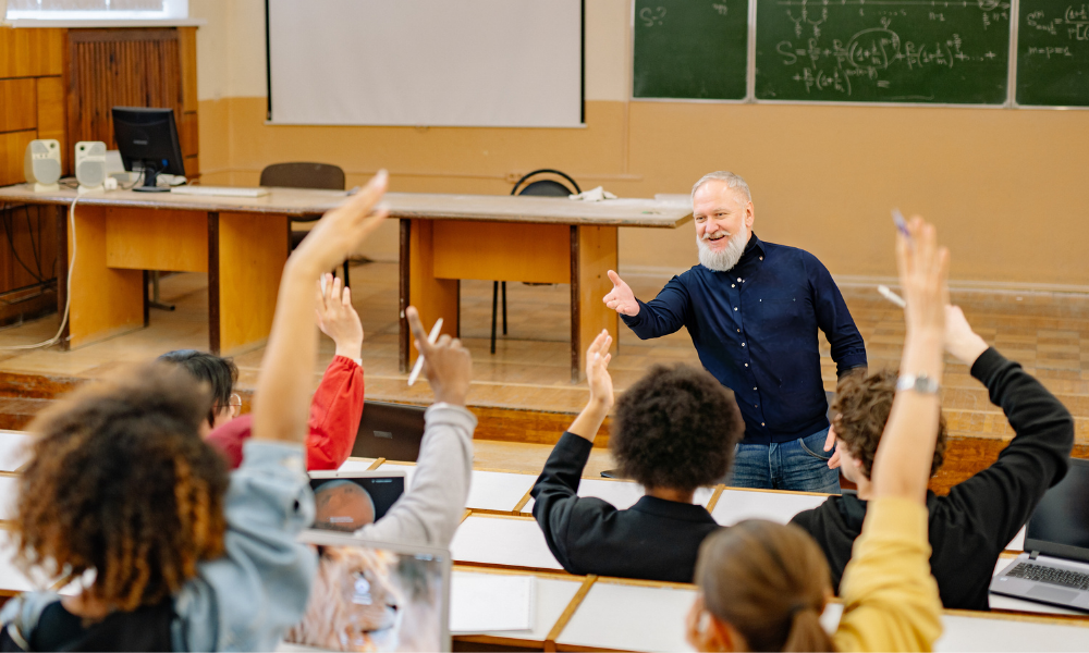 A professor at the front of the class while students raise their hands