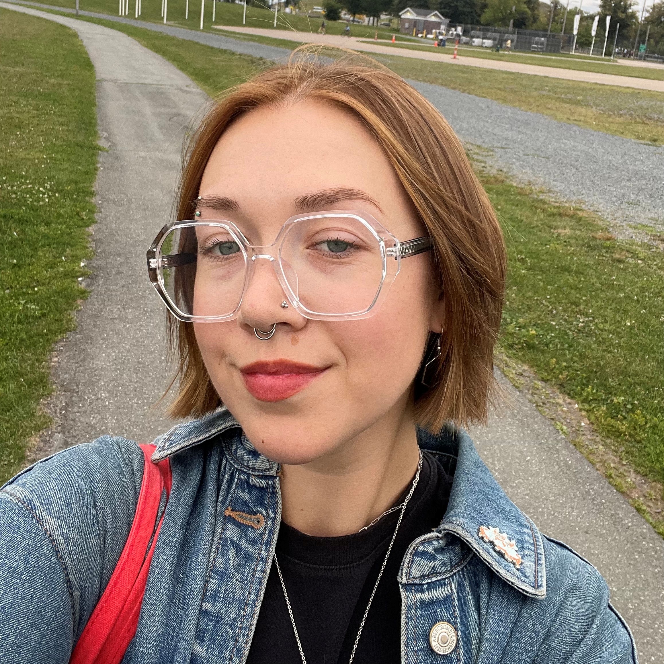 Photo of a woman from the shoulders up with short brownish-red hair and clear octagon glasses. She is grinning at the camera and has green-blue eyes. She is wearing a black shift, jean jacket and has a red purse on her shoulder against a gravel path and grass background.