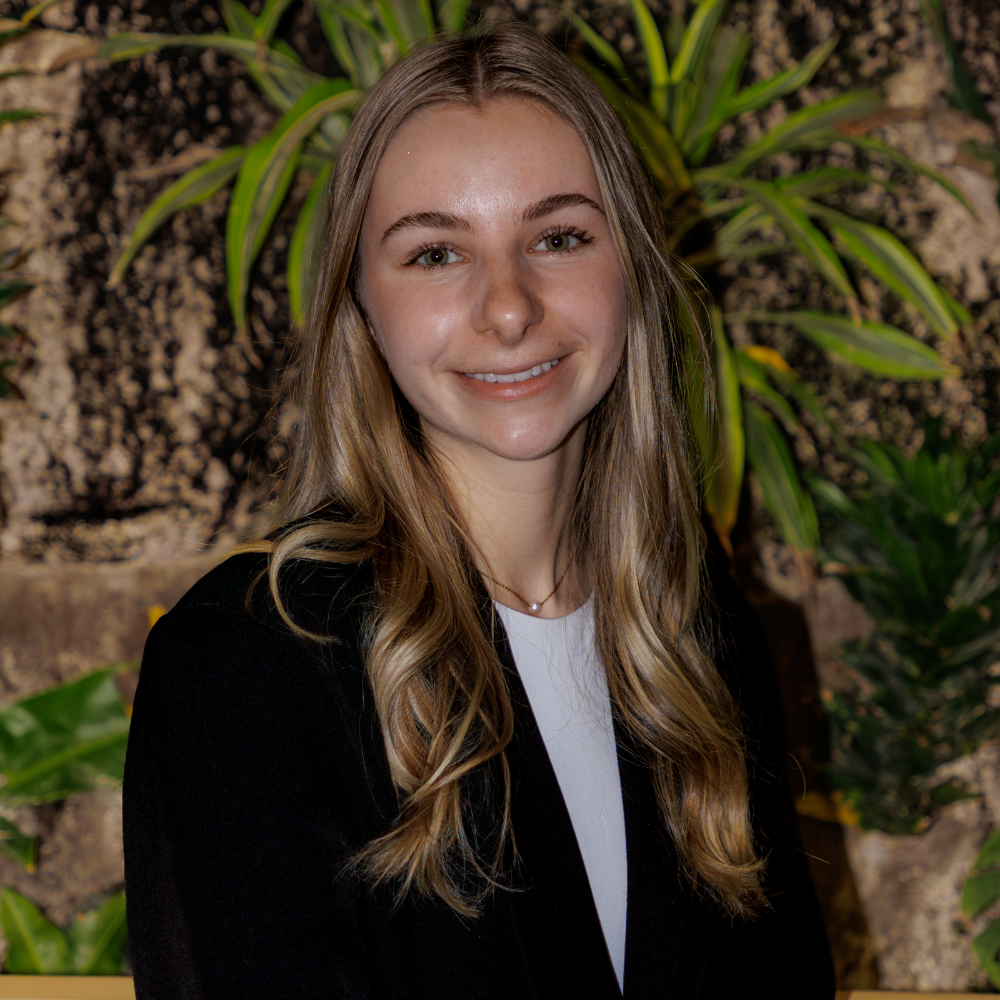 Sophia is a white woman with long blond hair. She wears a black blazer and stands in front of a green plant.
